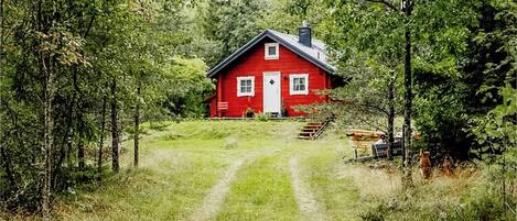 Una casa roja con puerta y ventanas blancas, rodeada de árboles y un sendero cubierto de césped que lleva a ella.