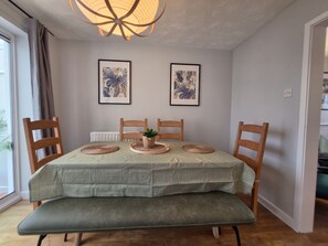 Un comedor con una mesa dispuesta para dos personas, dos cuadros con hojas tropicales en la pared y una lámpara de techo.