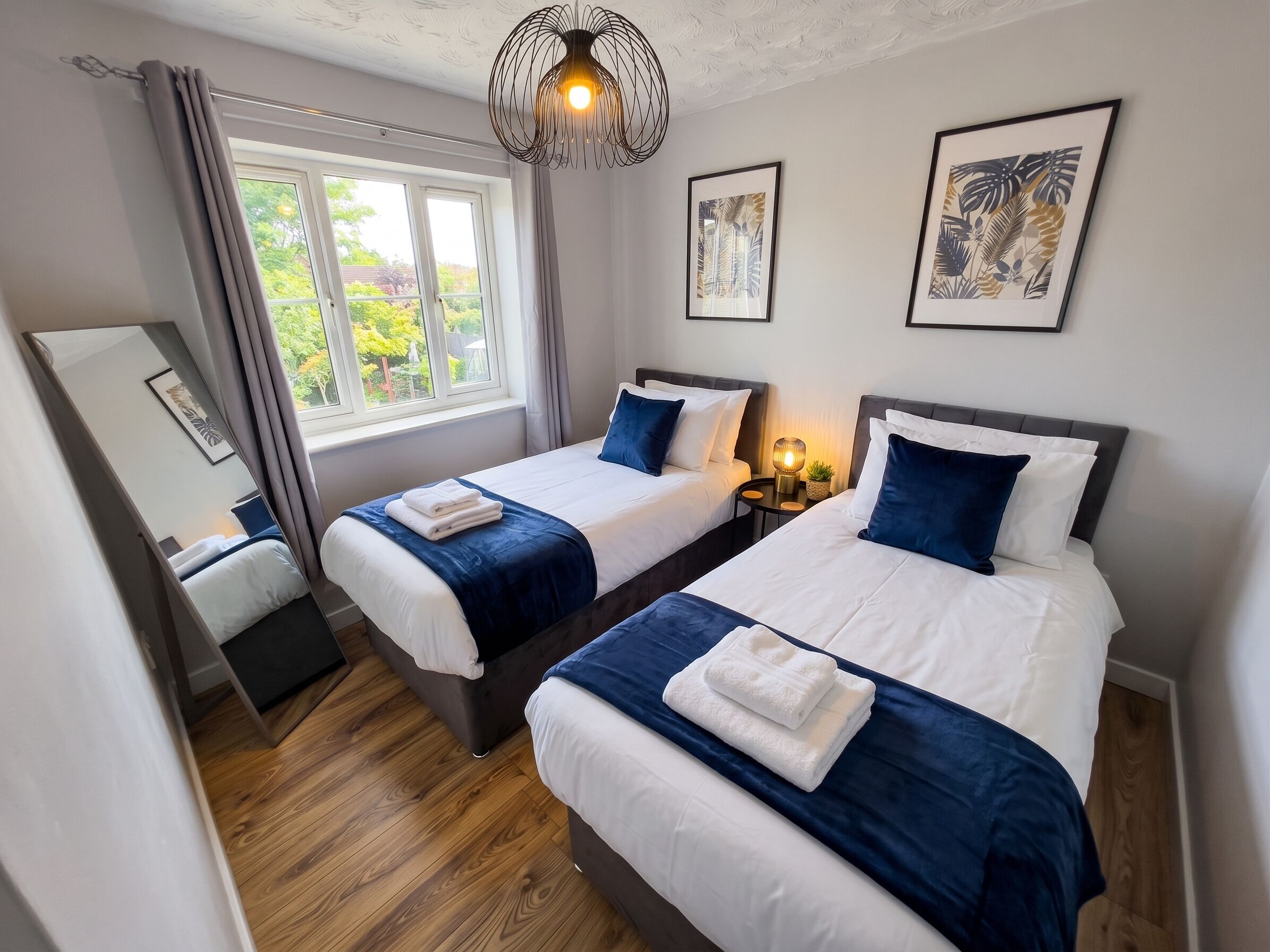 Une chambre d’hôtel avec deux lits, une fenêtre avec des rideaux, un miroir et des œuvres d’art encadrées au mur.
