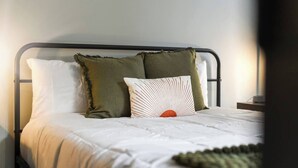 A single bed with a metal frame, white bedding, and three pillows of different textures and colors. - Cozy 1BR Downtown Pittsbu (Pittsburgh)