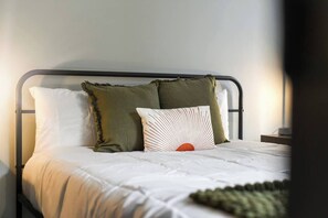 A single bed with a metal frame, white bedding, and three pillows of different textures and colors. - Cozy 1BR Downtown Pittsbu (Pittsburgh)