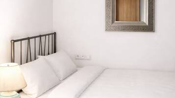 Una cama bien hecha con ropa de cama blanca, cabecera de metal negro, lámpara de noche y espejo de pared con marco decorativo.