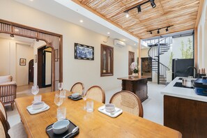 A dining area with a wooden table set for six, surrounded by wicker chairs. A kitchen area with a stove and sink is visible to the right. - Lily Hidden Homestay Spacious 150m² 2BR near Hoan Kiem Lake (Hanoi, Vietnam)