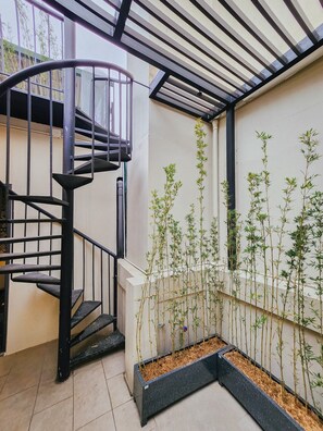 A spiral staircase with black railings and a metal handrail, leading to an upper level. There are tall bamboo plants in black rectangular planters along the wall. - Lily Hidden Homestay Spacious 150m² 2BR near Hoan Kiem Lake (Hanoi, Vietnam)