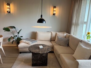A modern living room with a sectional sofa, a round marble coffee table, and a hanging pendant light. - Modern & cozy apartment in Capital area - scenic views in nature! (Garðabær)