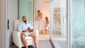 A man seated in a chair reading a book in a room with a glass wall, a woman and a child in the background, and a white door. - Fahana Canteras (LAS PALMAS DE GRAN CANARIA)