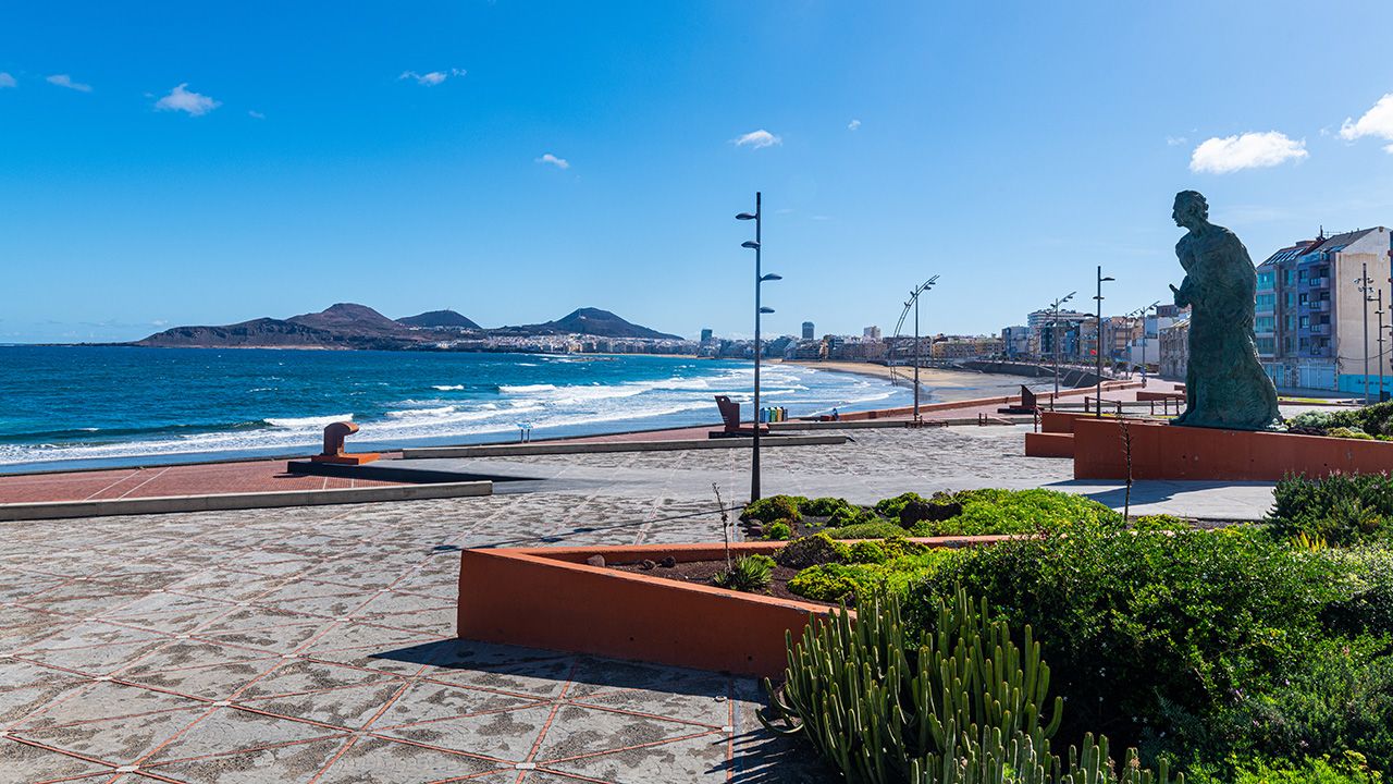 A coastal promenade with a statue, a clear sky, and a view of the sea and distant hills.