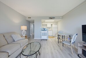 A compact living space with a sofa, a glass coffee table, a dining area with chairs, and a kitchenette. - Oceanfront 2BR/2BA Condo - Pool + Beach - Ormond-by-the-Sea (Ormond Beach)
