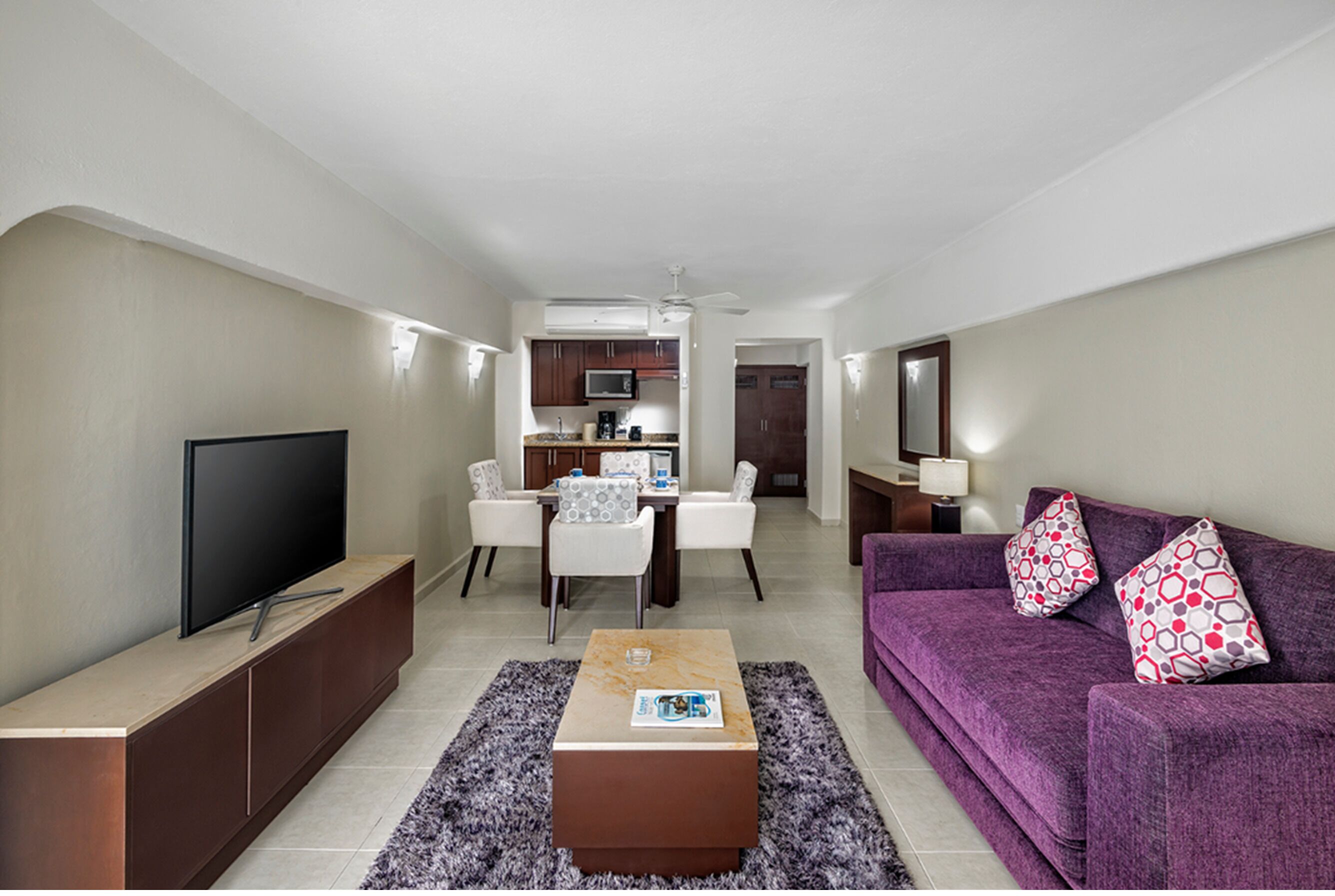A modern living room with a flat-screen TV, a coffee table, a purple sofa, and a dining area with chairs and a kitchen in the background.
