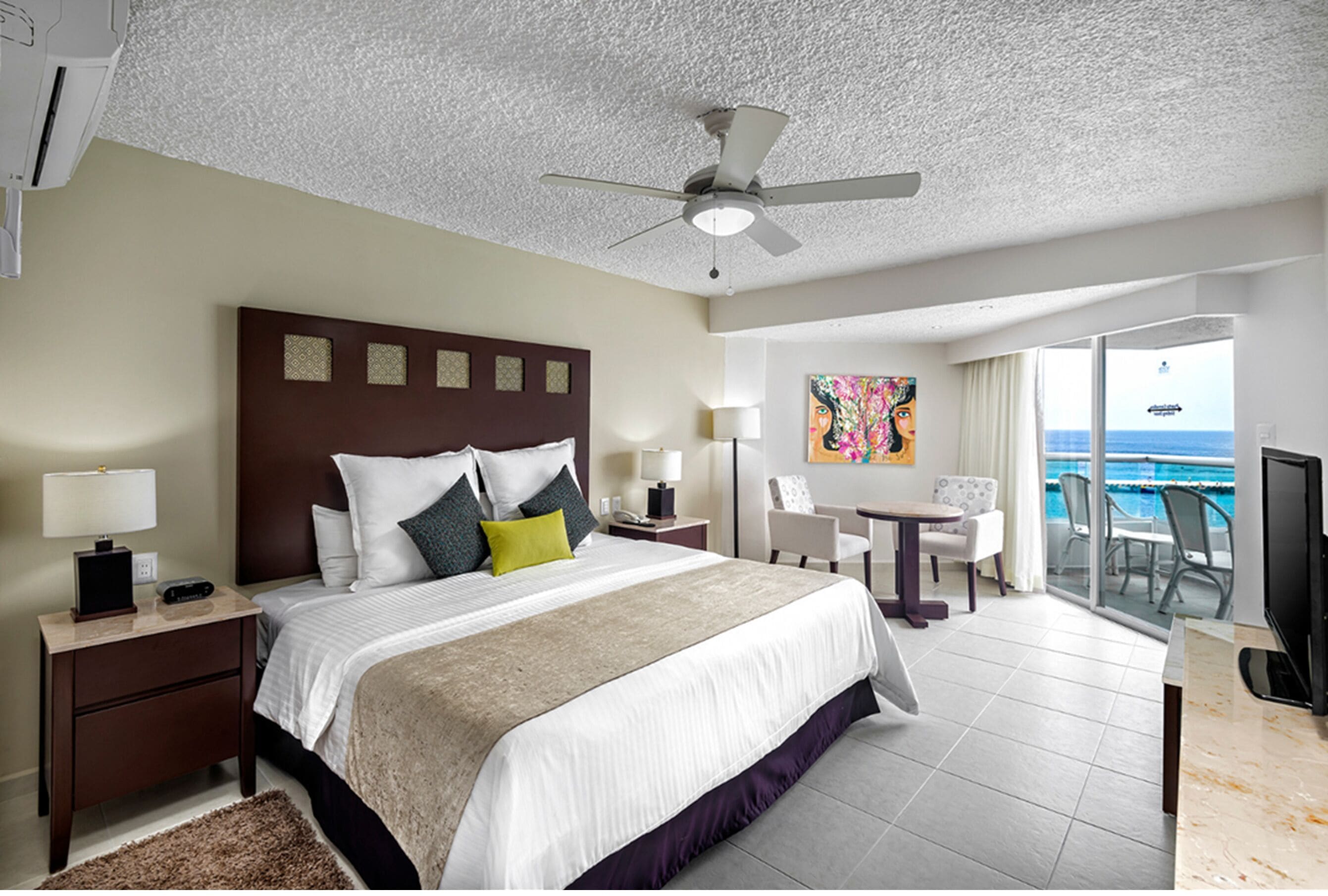 A hotel room with a large bed, a ceiling fan, a desk with a chair, a TV, and a balcony with a view of the ocean.