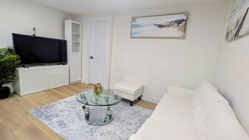 A living room with a flat-screen TV, a glass coffee table, a white sofa, and a painting of a beach scene.