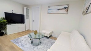A living room with a flat-screen TV, a glass coffee table, a white sofa, and a painting of a beach scene.