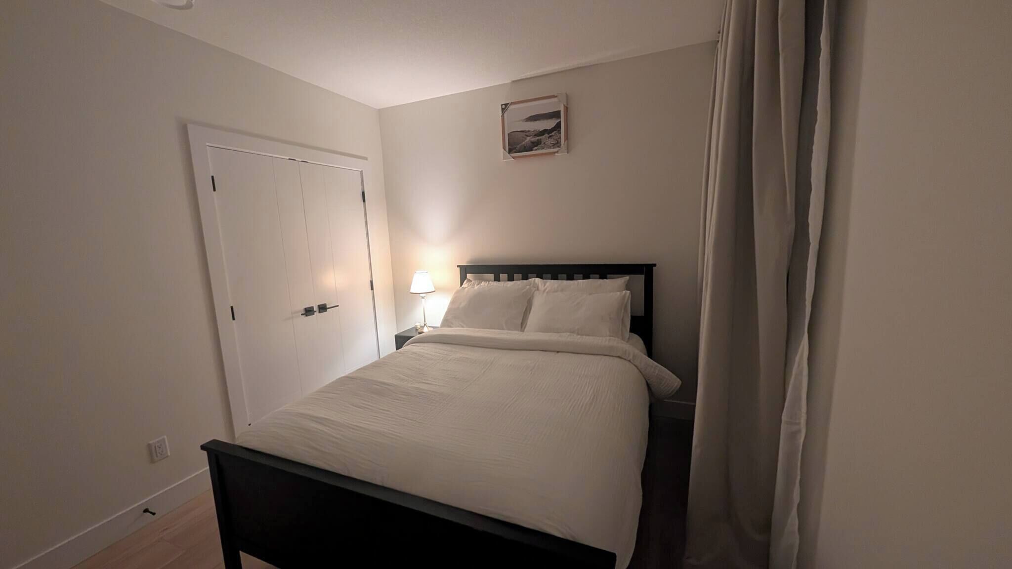 A bedroom with a bed, a nightstand, a lamp, a door, and a framed picture on the wall.