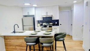 Cocina moderna con armarios blancos, refrigerador de acero inoxidable, horno doble y salpicadero de mármol. Hay tres taburetes de bar en la isla.
