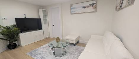 A modern living room with a white sofa, a glass coffee table, a flat-screen TV, and a painting on the wall.