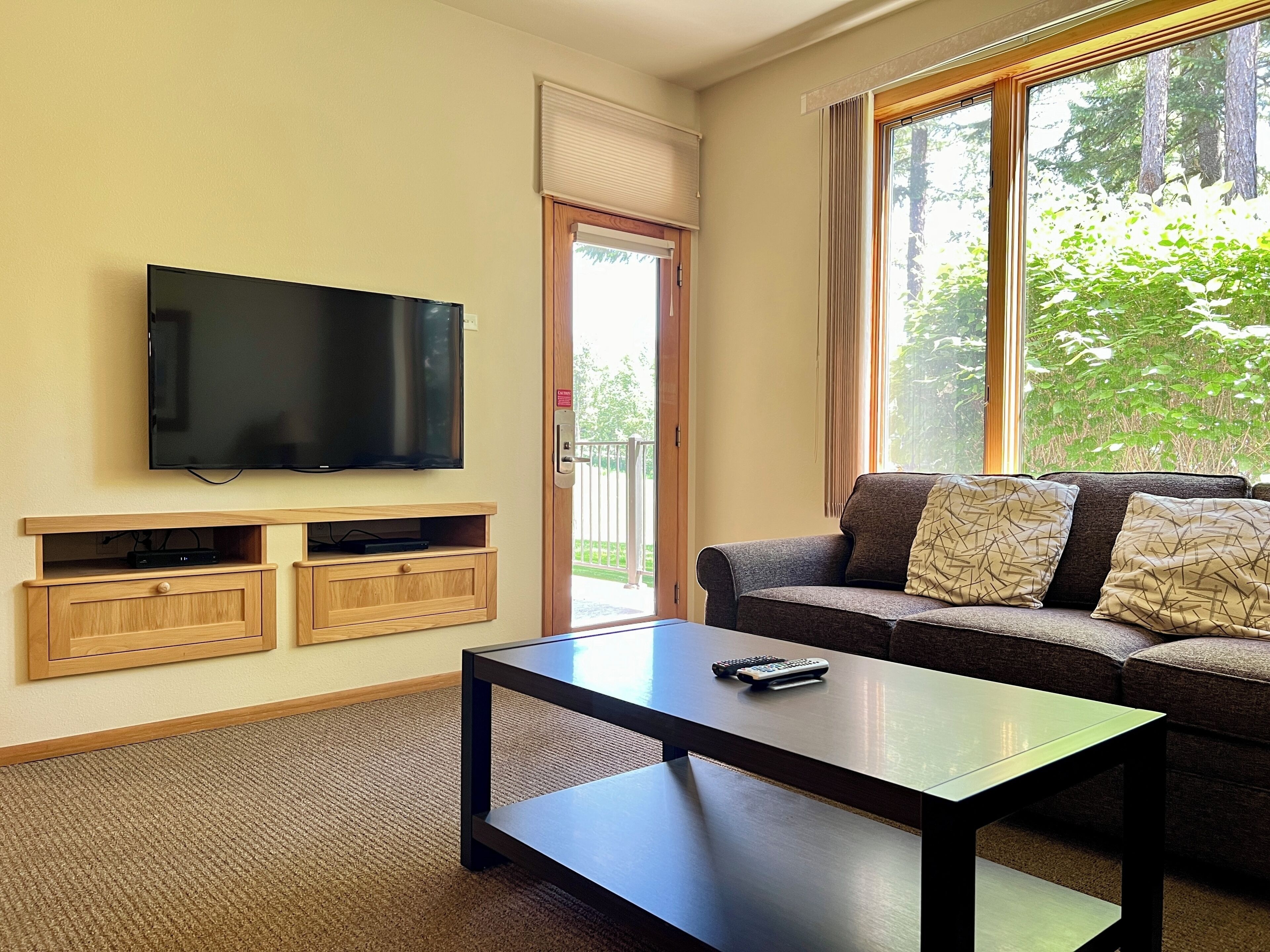 A living room with a flat-screen TV mounted on the wall, a sofa with patterned cushions, a coffee table, and a sliding glass door leading to an outdoor area.