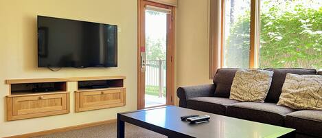 A living room with a flat-screen TV mounted on the wall, a sofa with patterned cushions, a coffee table, and a sliding glass door leading to an outdoor area.