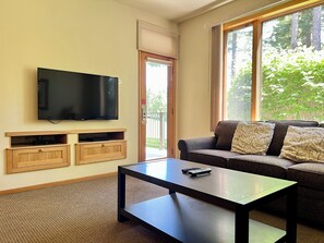 Ein Wohnzimmer mit an der Wand befestigtem Flachbildfernseher, einer Couch mit gemusterten Kissen, einem Couchtisch und einer Schiebeglastür zu einem Außenbereich.