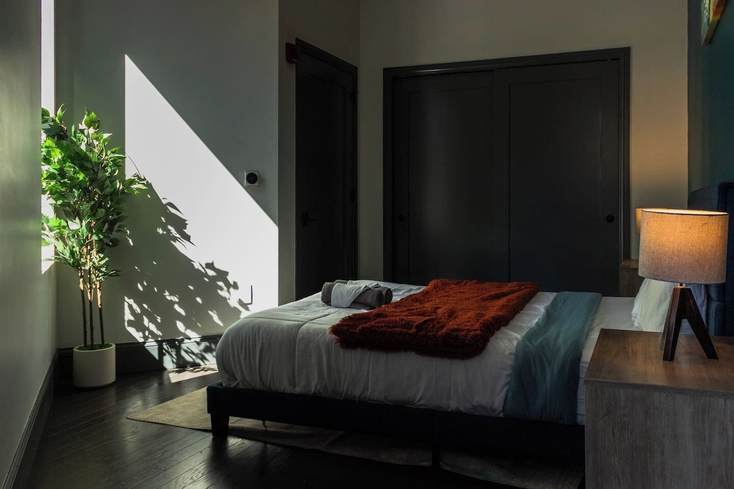 Un dormitorio con una cama, una mesita de noche, una lámpara, una planta en maceta y una puerta oscura.