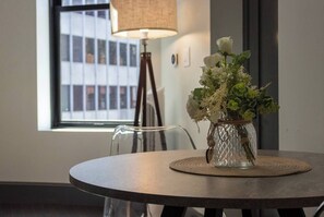 Ein runder Tisch mit einer klaren Glasvase, die weiße Blumen, eine Lampe und einen Stuhl enthält, in einem Zimmer mit einem Fenster und einem an der Wand befestigten Lichtschalter.