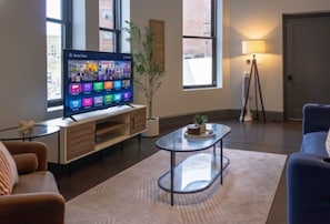 Ein modernes Wohnzimmer mit Fernseher auf Holzfuß, einem Glastisch, einem blauen Sofa und einer Lampe.