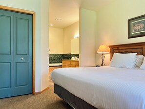 A hotel room with a bed, a nightstand, a lamp, a bathroom with a mirror, and a door. - Meadow Lake Private Timeshares (Columbia Falls)