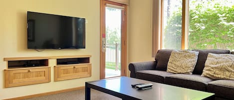 A living room with a flat-screen TV mounted on the wall, a sofa with patterned cushions, a coffee table, and a sliding glass door leading to an outdoor area.