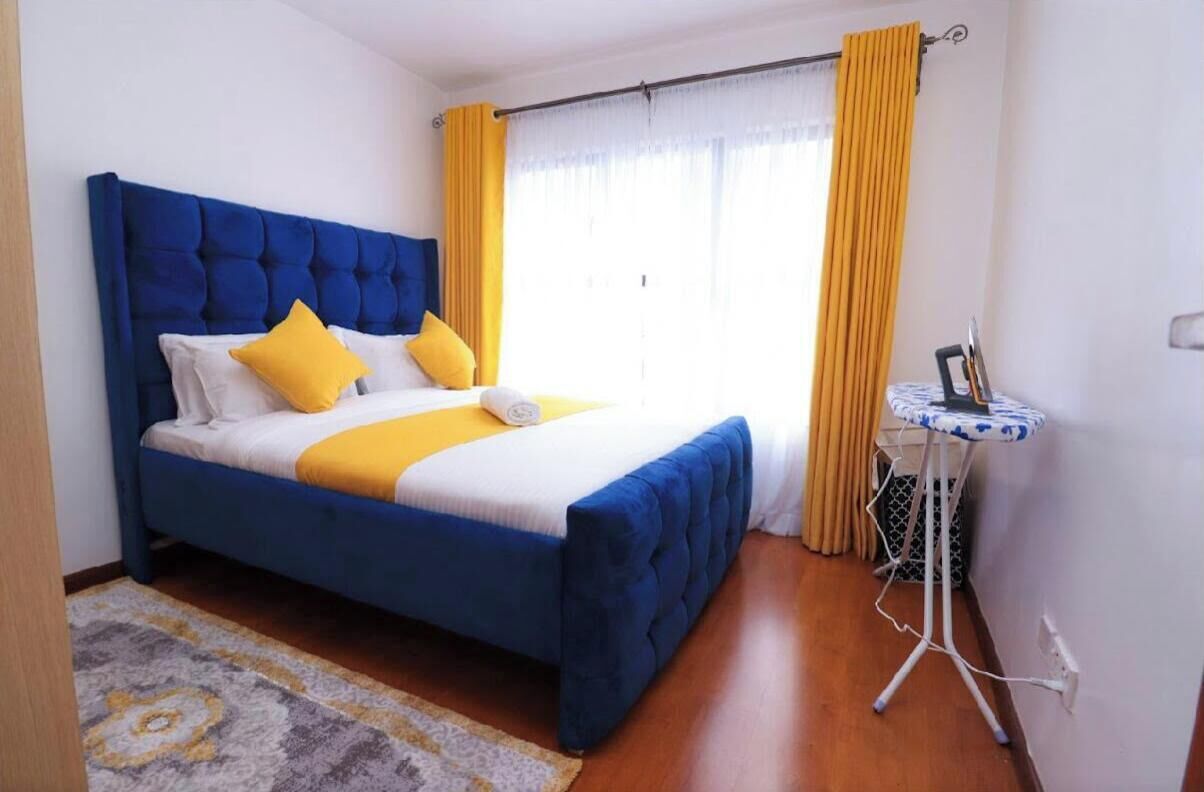 Un dormitorio con cama azul, detalles en amarillo, una mesita redonda y una ventana con cortinas.