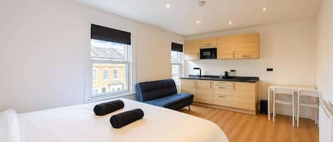 A compact bedroom with a bed, a small seating area, a kitchenette, and a window with a view.