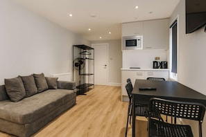 A modern living room with a sofa, a dining table with chairs, a kitchen area with a microwave, and a black metal shelf unit. - Large Studio | Private Bathroom & Kitchenette NW10 (Greater London)