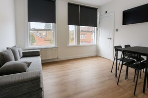 Ein Zimmer mit einer Couch, einem Esstisch mit Stühlen und zwei Fenstern mit Rollos.