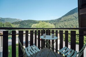 A balcony with a view of a forested valley, a small table with two cups, and a bench. - Napoléon - Cosy 80m² Duplex | Central | Sleeps 6 | Parking (Les Houches)