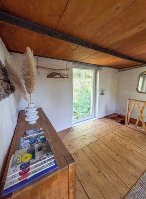 A room with wooden flooring, a wooden desk with a vase and magazines, a window with a view of greenery, and a mirror on the wall.