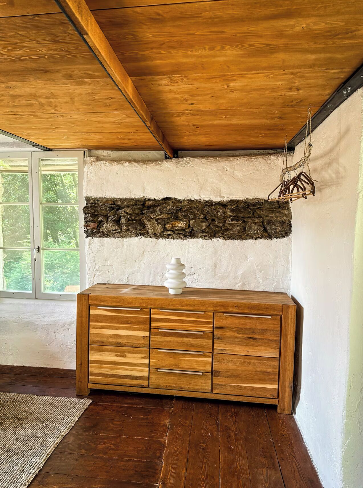 A wooden cabinet with drawers, a stone wall, and a hanging light fixture in a room with wooden flooring and a window.