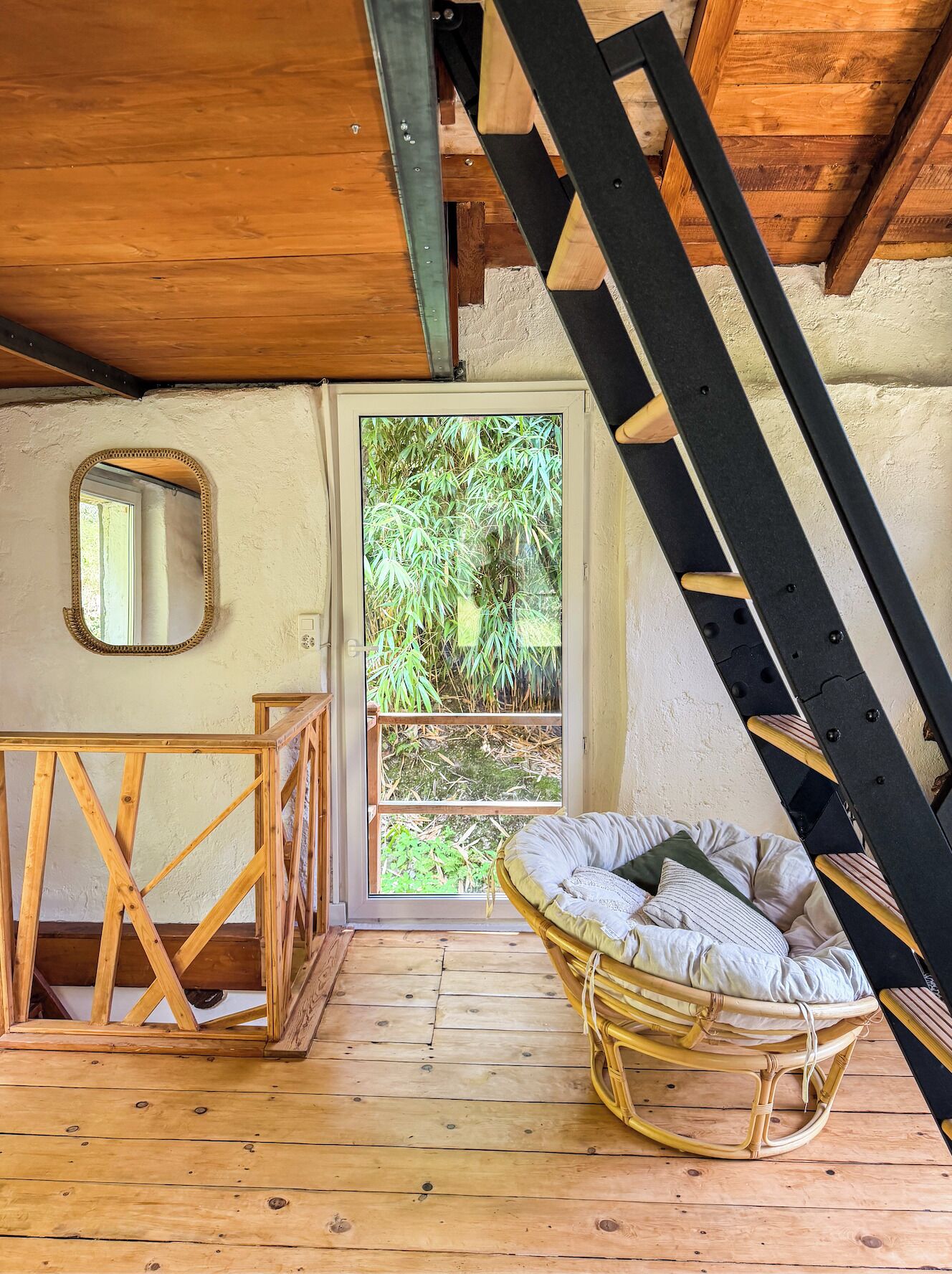 Een houten interieur met een schuine kap, een rieten stoel, een houten tafel en een deur naar een tuin.