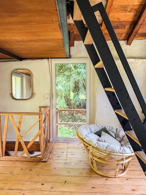 A wooden interior with a sloped ceiling, a wicker chair, a wooden table, and a door leading to a garden.