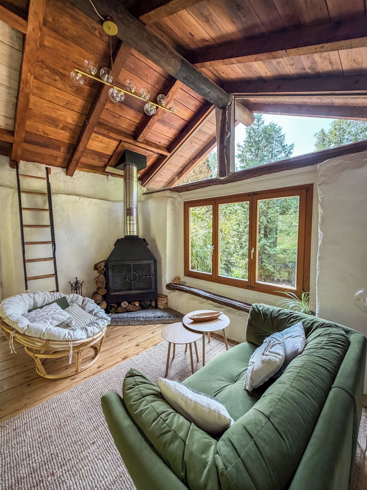 Ein gemütliches Zimmer mit einem grünen Sofa, einem Korbstuhl, einem kleinen Tisch, einem Kamin und einem Fenster mit Blick auf Bäume.
