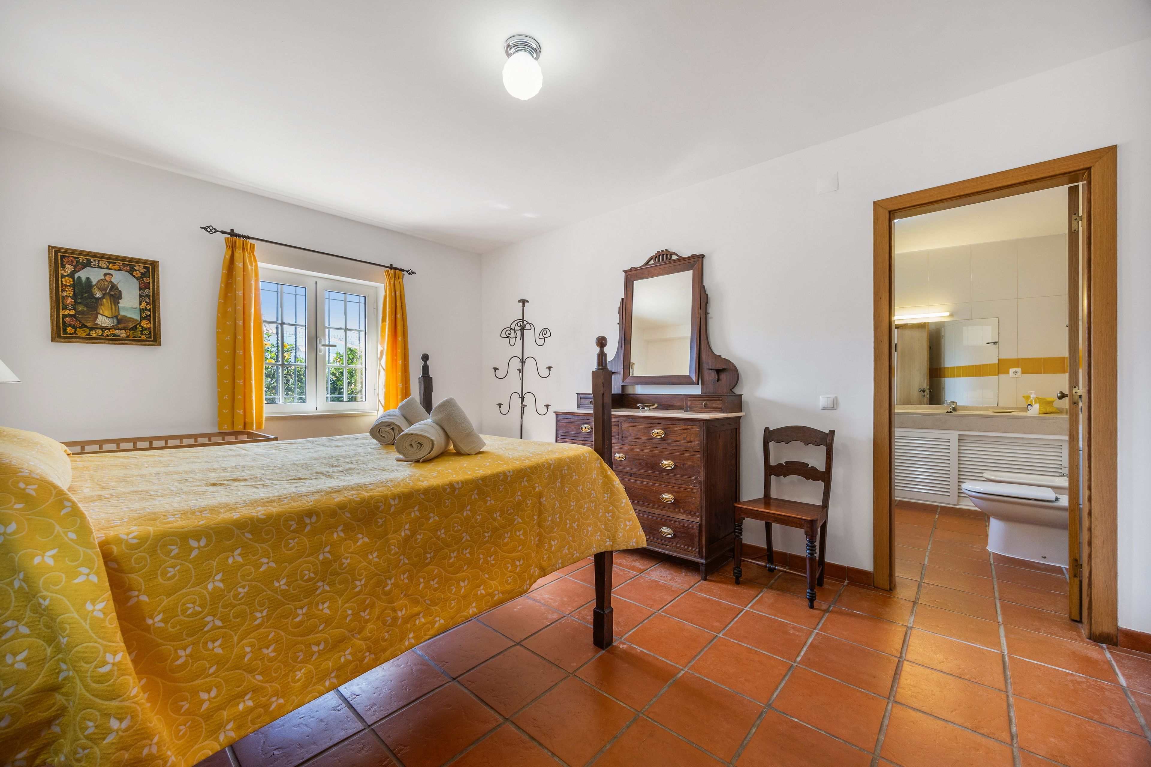 Une chambre à coucher avec un lit, une commode, une chaise, un miroir et une fenêtre avec des rideaux.