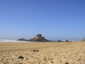 Ein Sandstrand mit einer markanten Felsformation in der Ferne und einem strahlend blauen Himmel.