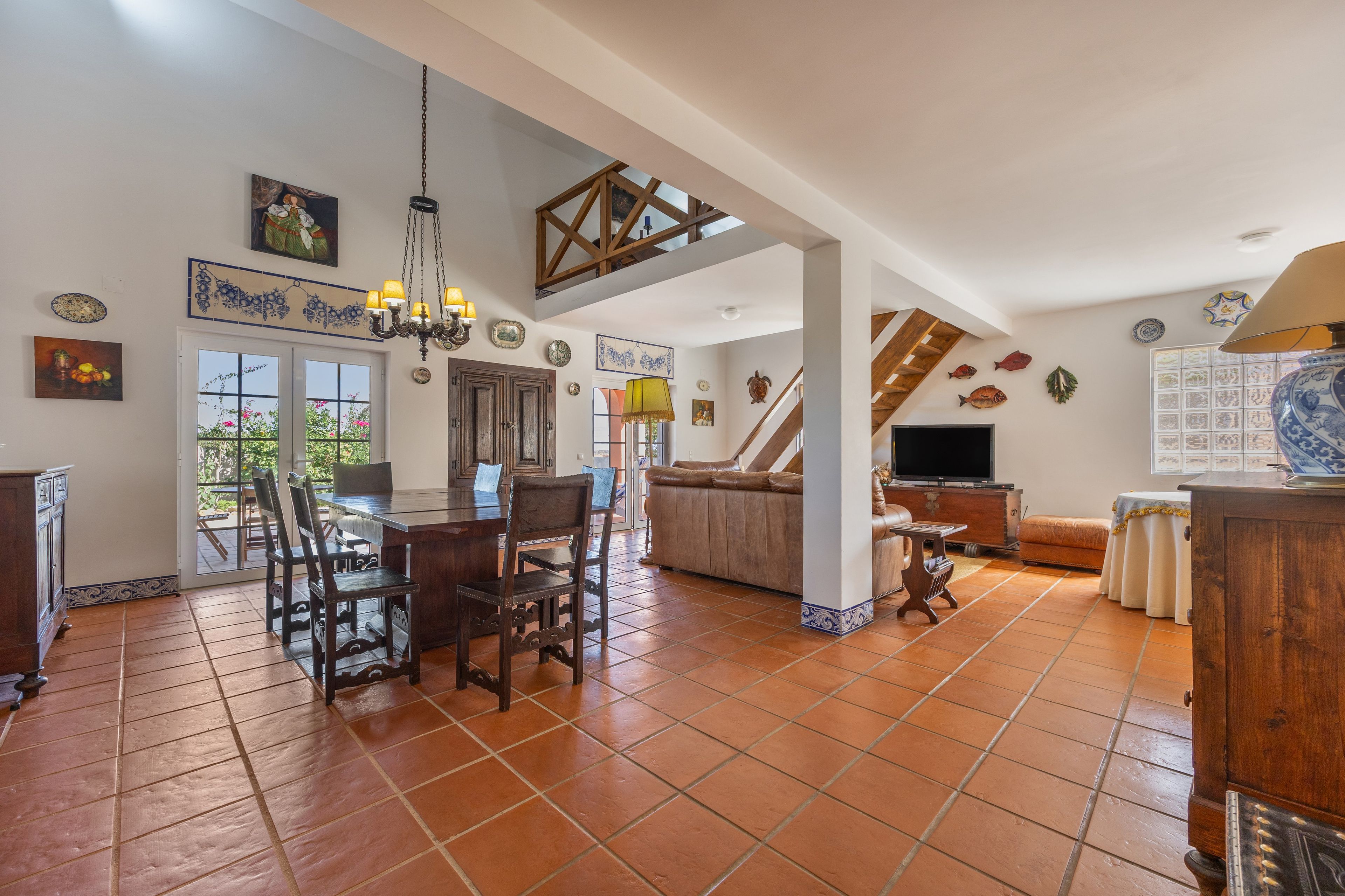 A spacious living area with a dining table, chairs, a sofa, and a TV. There is a staircase leading to an upper level.