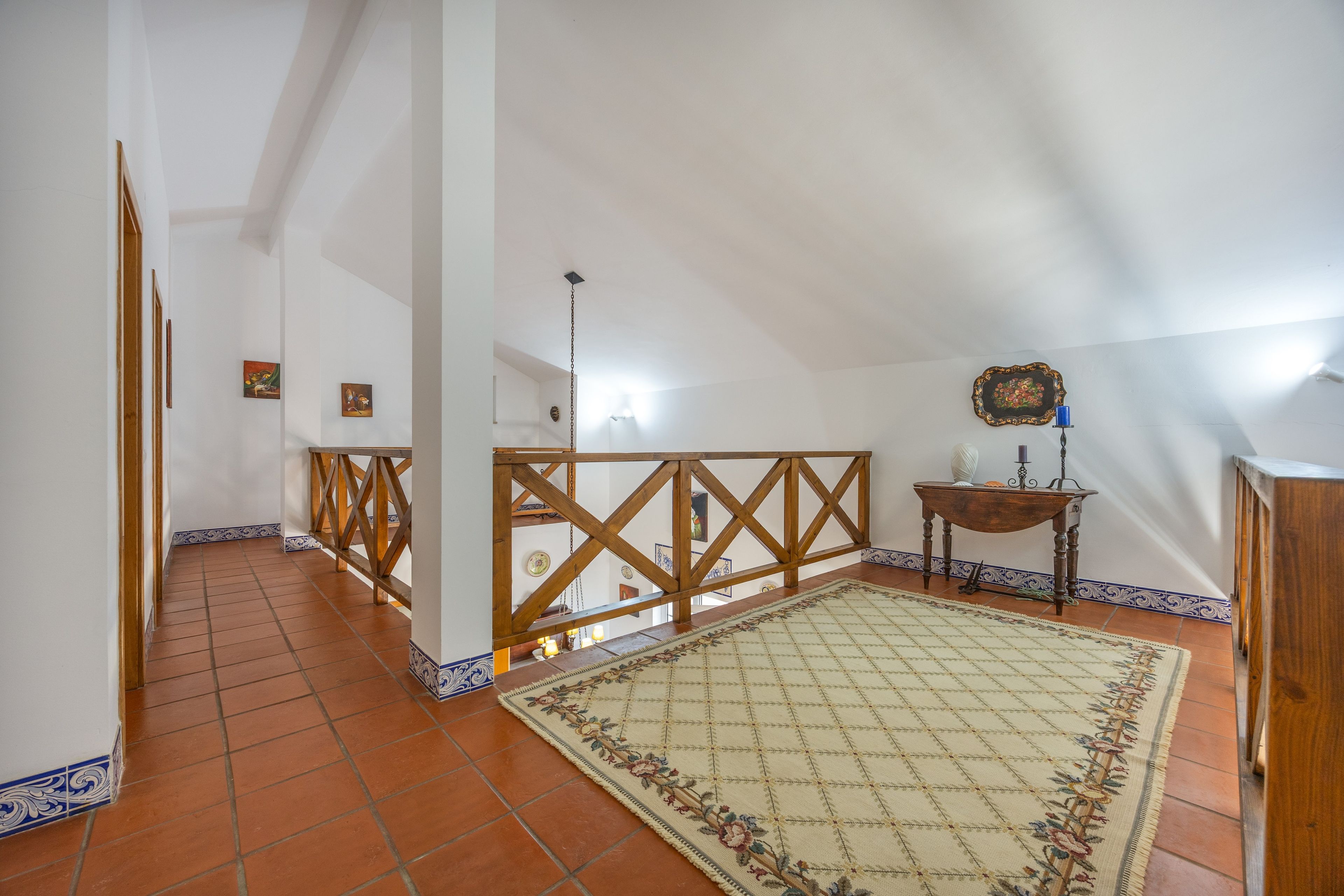 Une pièce mansardée spacieuse avec un plafond en pente, une rambarde en bois, un tapis à motifs et une petite table sur laquelle est posée une bougie.