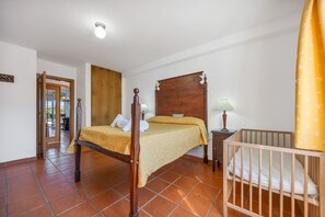 A bedroom with a wooden four-poster bed, a nightstand with a lamp, a crib, and a bathroom visible through an open door.