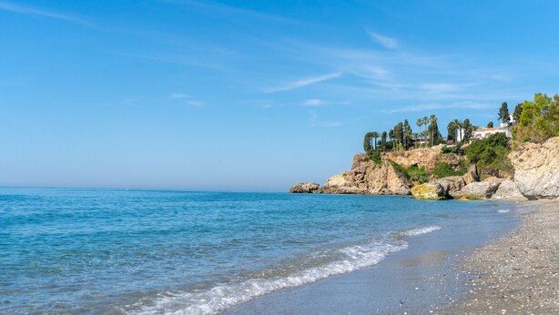 Uma praia com águas azul-claras, areal pedregoso e falésias rochosas com vegetação.