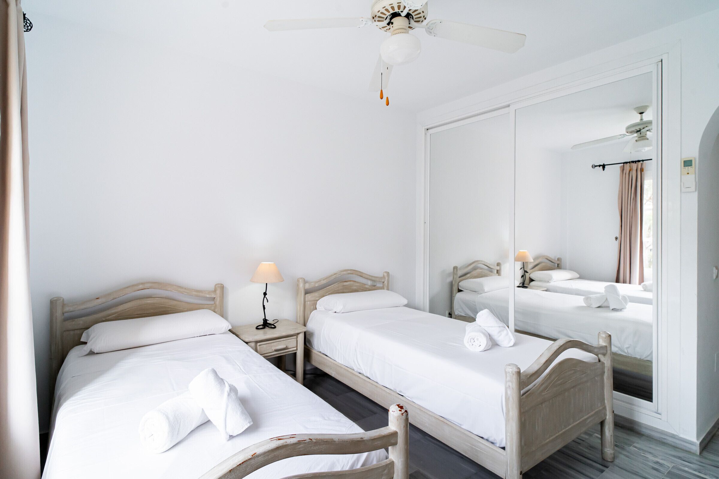 A hotel room with two single beds, a ceiling fan, a bedside table with a lamp, and a large mirror reflecting the room.