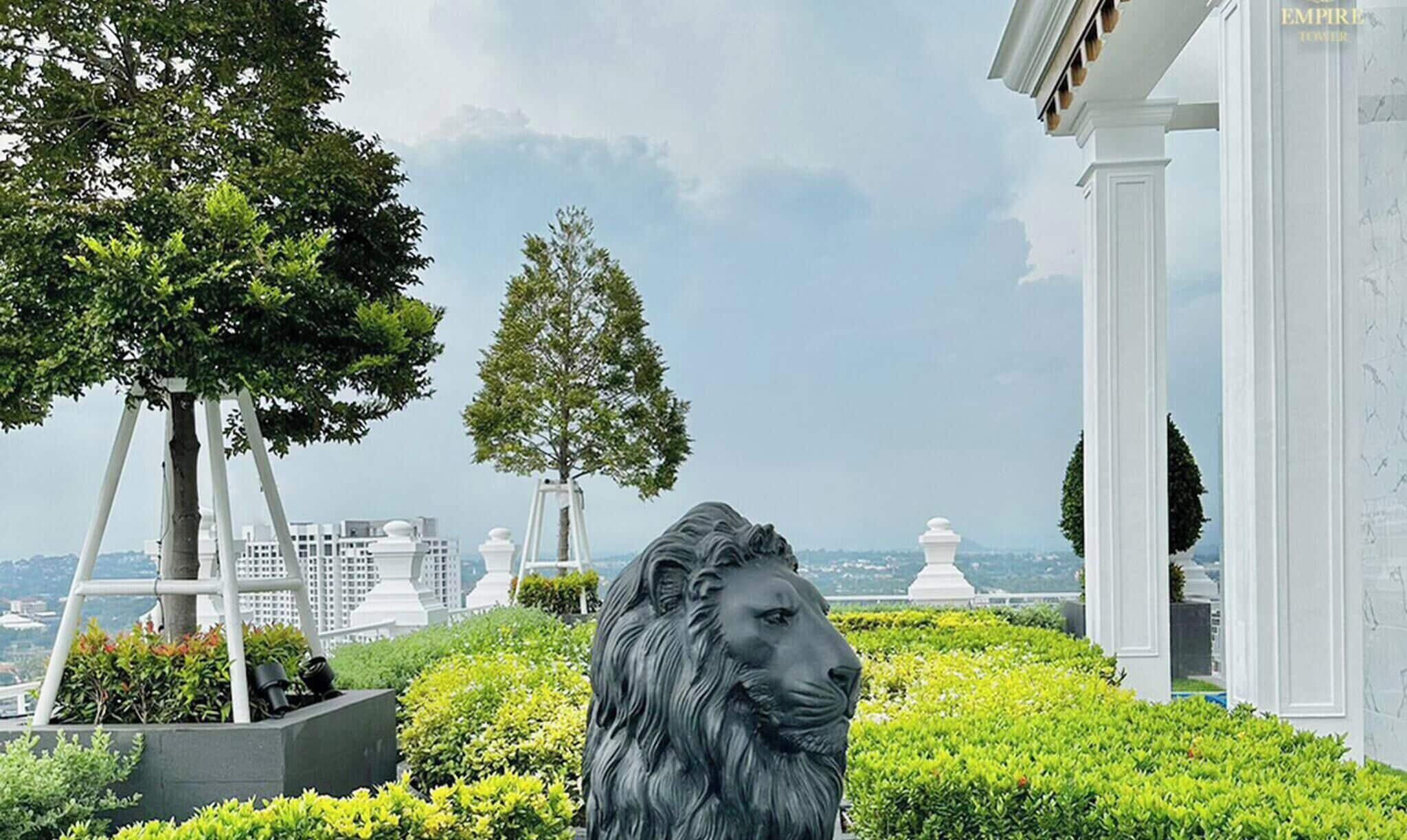 Una estatua de león en un área ajardinada con árboles y una vista de edificios al fondo.