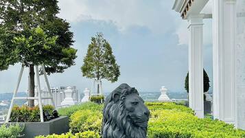 Una estatua de león en un área ajardinada con árboles y una vista de edificios al fondo.