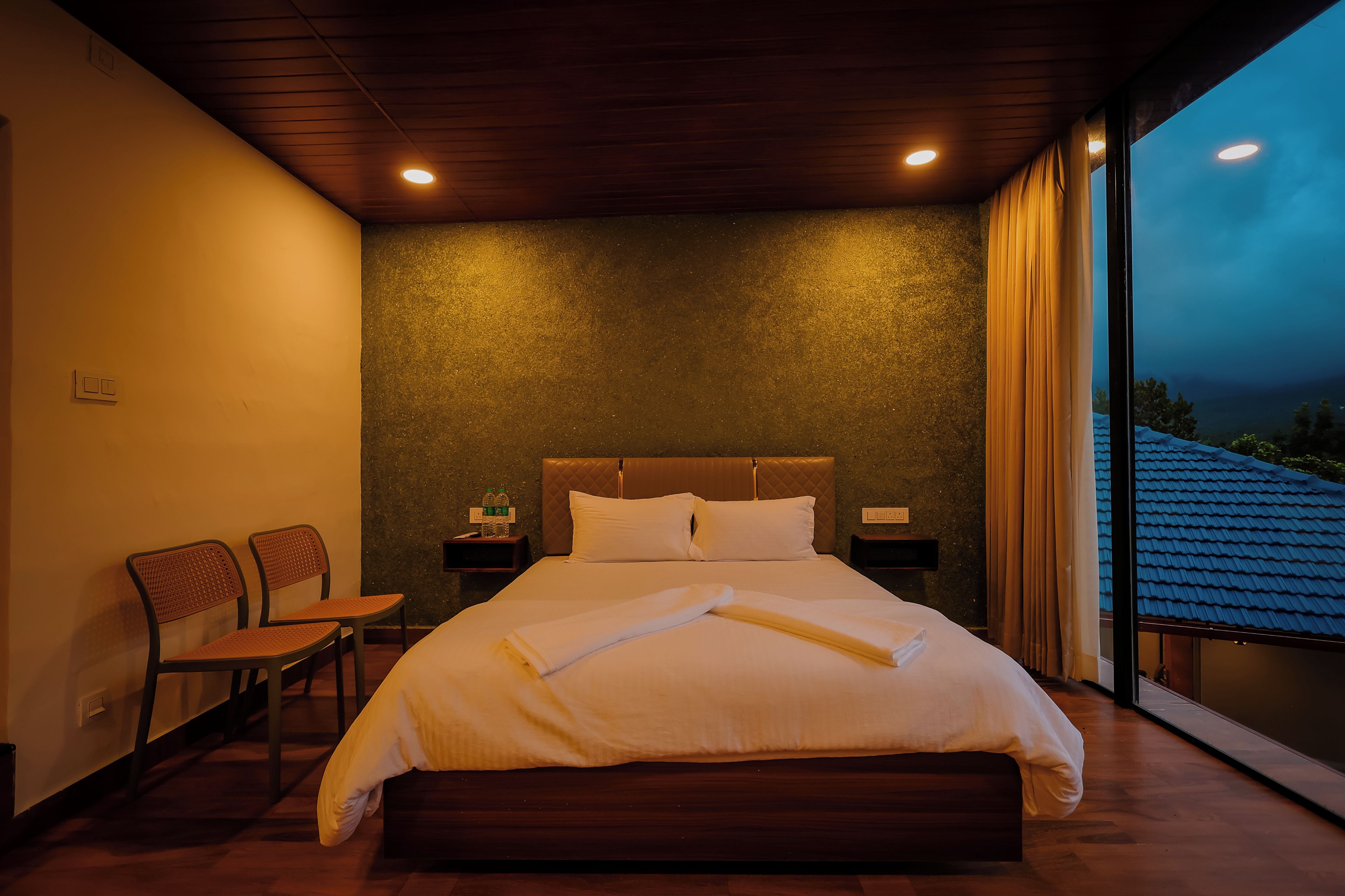 Habitación de hotel con una cama grande, dos mesitas de noche, una silla y vistas al exterior.