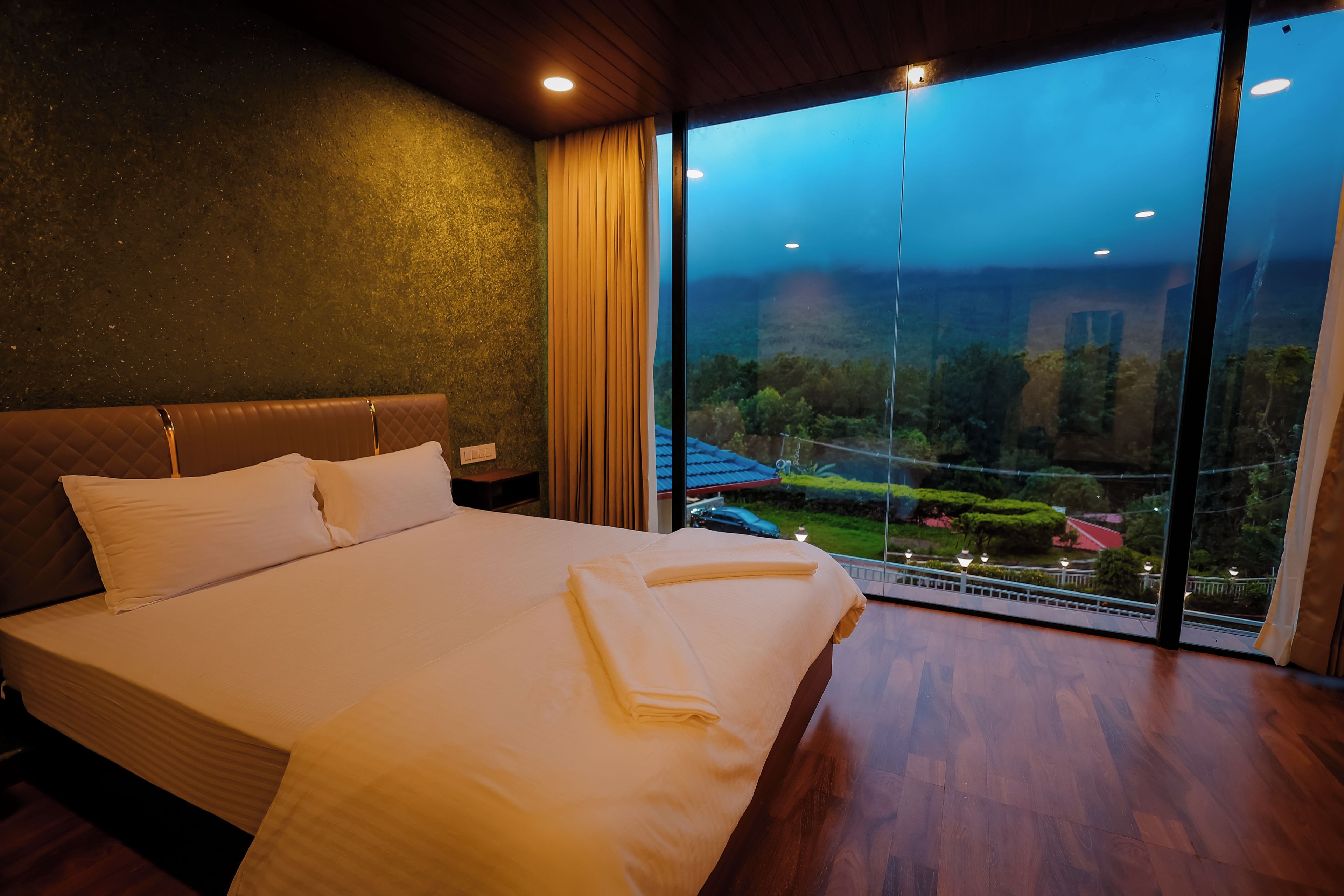 A hotel room with a large bed, a view of a garden through floor-to-ceiling windows, and a wooden ceiling.