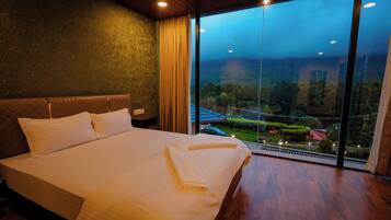 A hotel room with a large bed, a view of a garden through floor-to-ceiling windows, and a wooden ceiling.