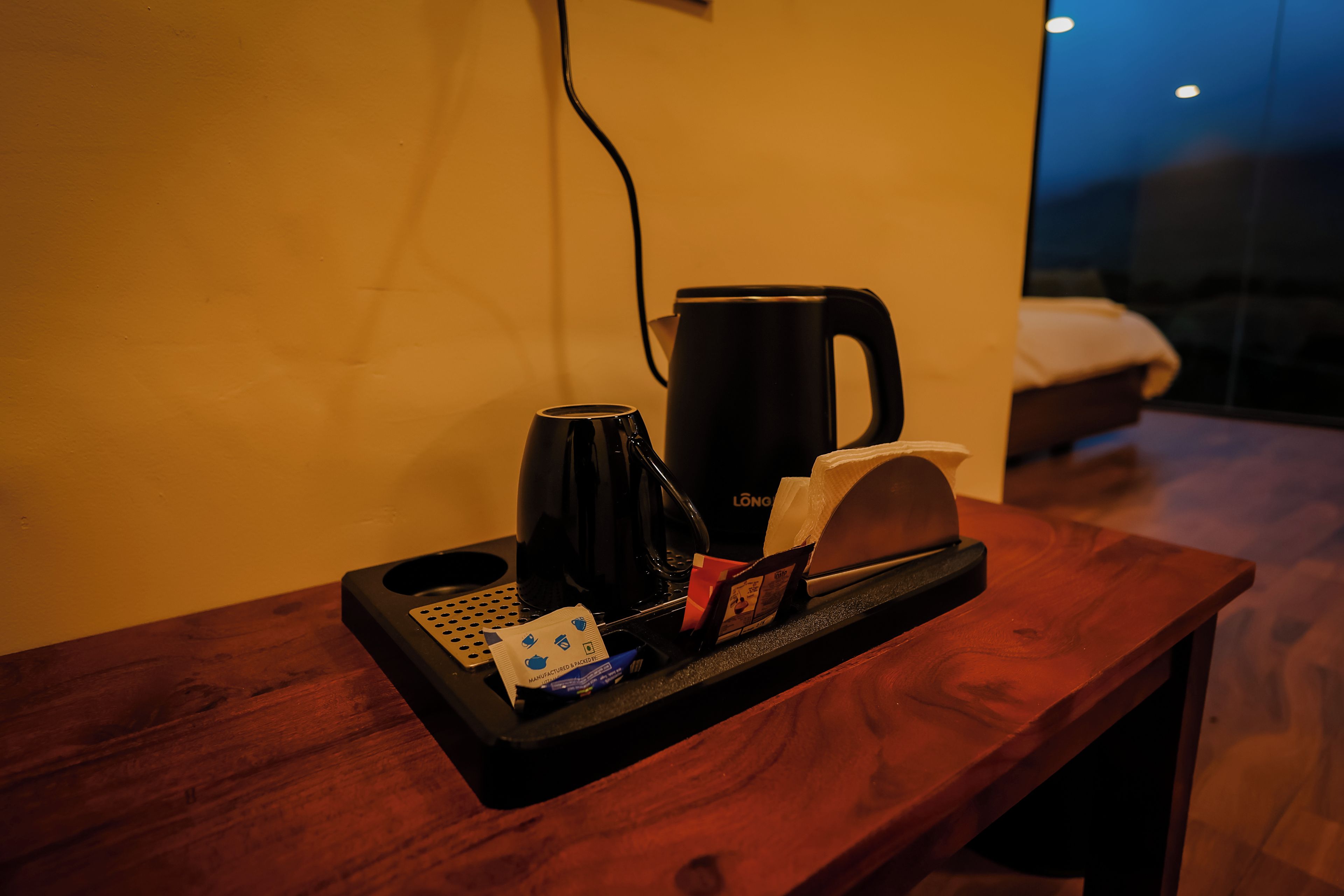 Una habitación de hotel con una mesita de noche de madera, una cafetera negra, una bandeja con un rollo de pañuelos, un paquete de té y un control remoto.